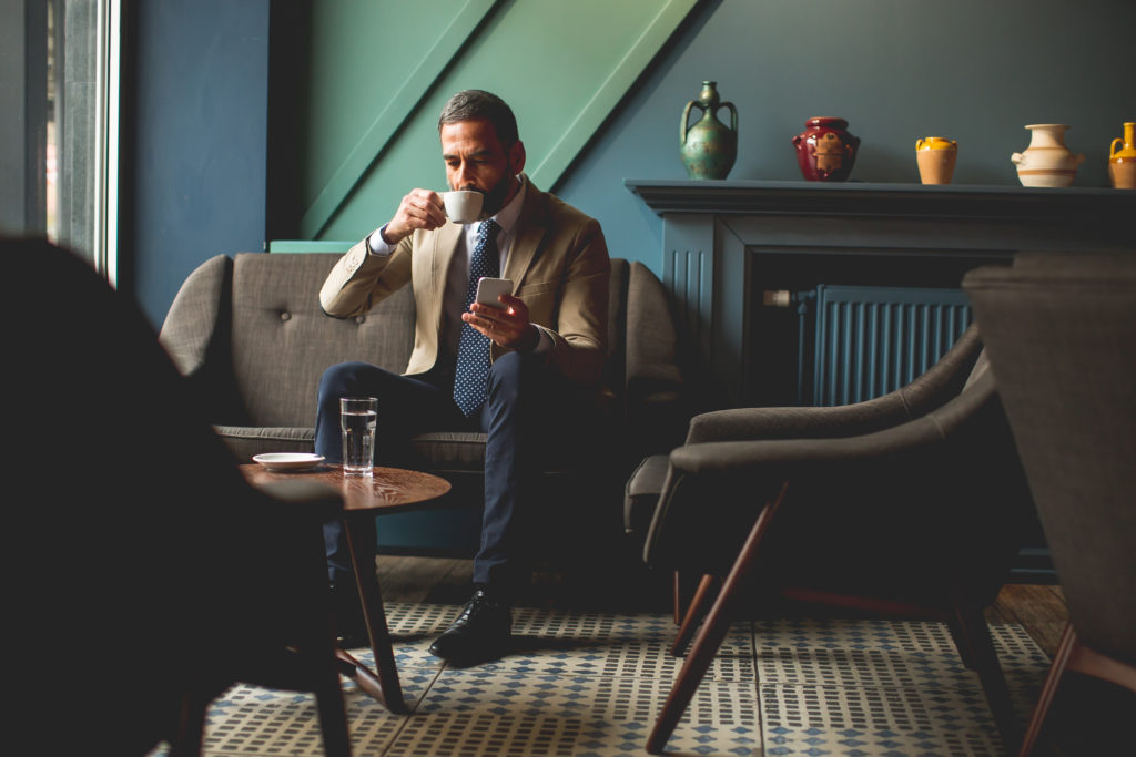 View at middle-aged businessman drinking coffee and using mobile phone in the loby